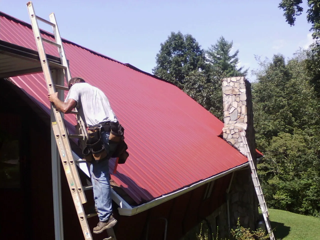 Roofer on a ladder beside a red metal roof and stone chimney — Soffit & Fascia Repair by Roofing Brothers in Boys Town